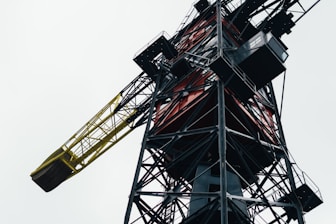 A close-up view of an industrial crane showing its intricate metal structure, including a yellow and black jib. It appears to be a large, robust piece of machinery, with various components and platforms.