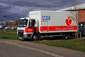 A large white and red NHS truck is parked on a grassy area next to a sidewalk. The sides of the truck display the message 'Please give blood' along with the NHS logo and contact information. Nearby, other vehicles are parked, and a few trees and buildings are visible under a cloudy sky.