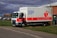 A large white and red NHS truck is parked on a grassy area next to a sidewalk. The sides of the truck display the message 'Please give blood' along with the NHS logo and contact information. Nearby, other vehicles are parked, and a few trees and buildings are visible under a cloudy sky.
