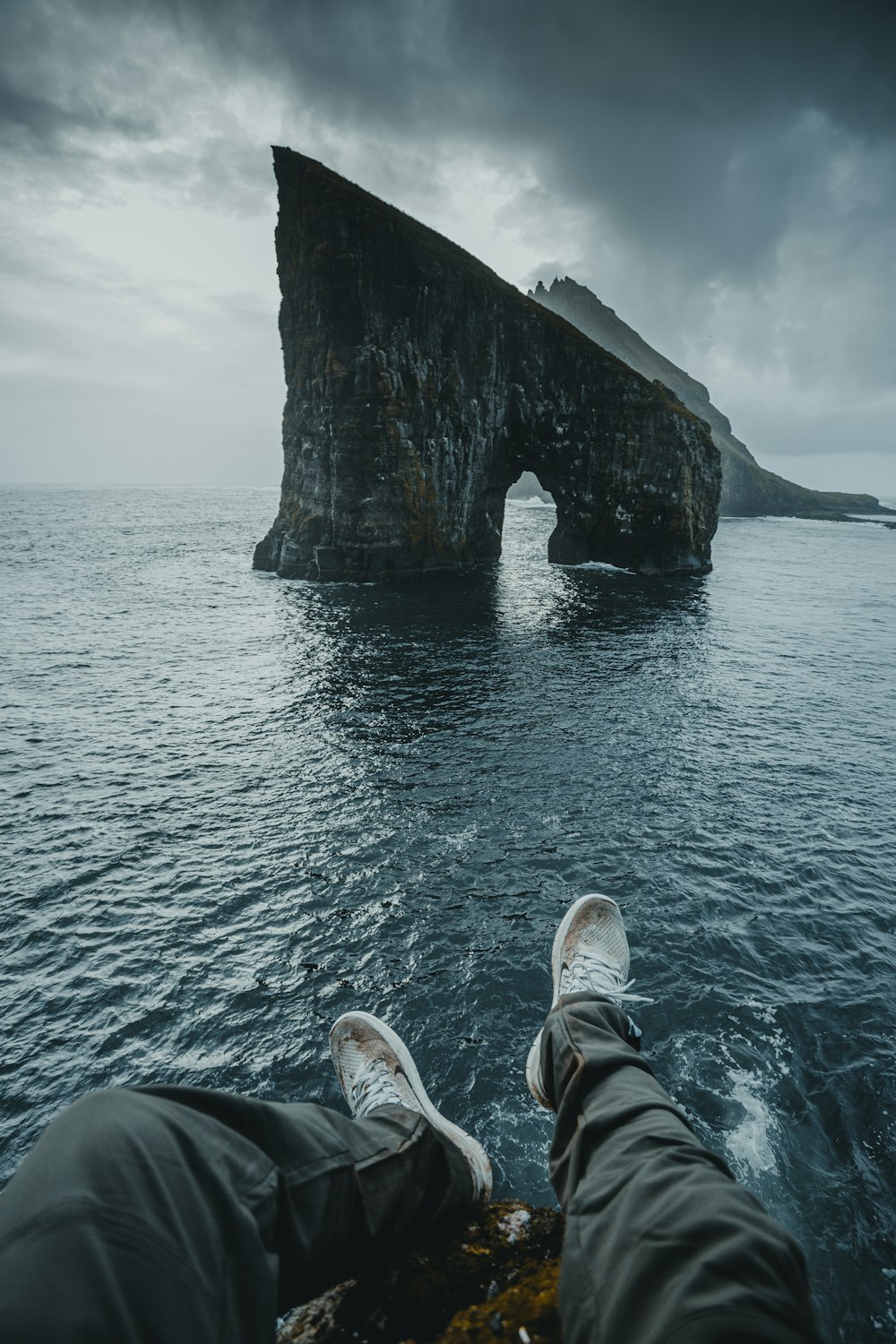 person in gray pants and white sneakers sitting on rock formation in front of sea during