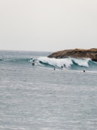 Several surfers ride along a series of rolling waves with a rocky outcrop visible in the background. The water has a cool, subdued tone, and the waves create dynamic movement across the image.