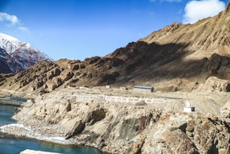 A rugged mountainous landscape with rocky terrain dominates the scene. A river curves alongside the base of the mountains, reflecting the clear blue sky. Snow-capped peaks are visible in the distance. A small facility or building sits on a plateau midway up the slope, and there are man-made religious structures such as stupas visible on the mountainside.