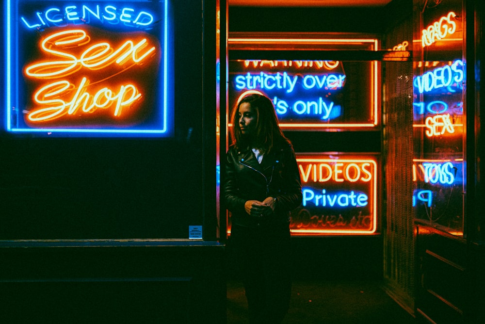 woman in black jacket standing in front of blue and red UNKs neon signage