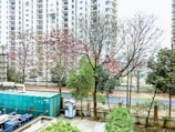 A row of tall residential buildings serves as the backdrop, with partially bare trees in the foreground showing traces of red leaves. A large green industrial unit is visible alongside a small security booth, all bordered by a fenced garden area.