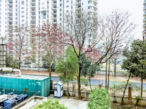 A row of tall residential buildings serves as the backdrop, with partially bare trees in the foreground showing traces of red leaves. A large green industrial unit is visible alongside a small security booth, all bordered by a fenced garden area.