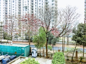 A row of tall residential buildings serves as the backdrop, with partially bare trees in the foreground showing traces of red leaves. A large green industrial unit is visible alongside a small security booth, all bordered by a fenced garden area.