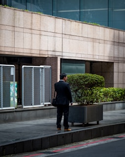 A man in a suit stands on the sidewalk next to a large planter with a shrub. The setting appears to be an urban area, with buildings featuring glass windows and a brick facade. It seems to be a quiet moment, possibly in the early morning or late afternoon.