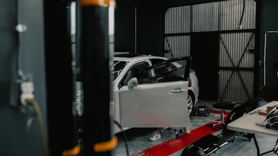 A workshop featuring a white car with its driver's side door open. Various tools and equipment are scattered around, including a stool, a table with items on it, and a black cylindrical object in the foreground.