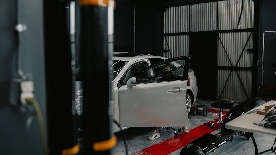A workshop featuring a white car with its driver's side door open. Various tools and equipment are scattered around, including a stool, a table with items on it, and a black cylindrical object in the foreground.