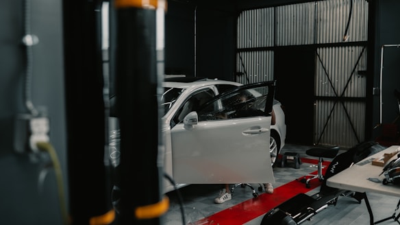 A workshop featuring a white car with its driver's side door open. Various tools and equipment are scattered around, including a stool, a table with items on it, and a black cylindrical object in the foreground.
