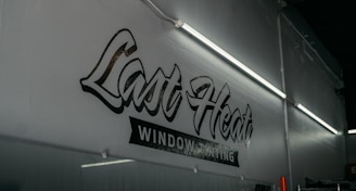 A dimly lit interior featuring a wall with stylized text that reads 'Last Heat Window Tinting.' The wall is adorned with fluorescent lights that cast a soft glow over the area.