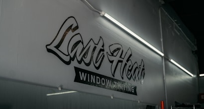 A dimly lit interior featuring a wall with stylized text that reads 'Last Heat Window Tinting.' The wall is adorned with fluorescent lights that cast a soft glow over the area.