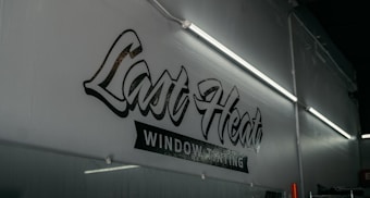 A dimly lit interior featuring a wall with stylized text that reads 'Last Heat Window Tinting.' The wall is adorned with fluorescent lights that cast a soft glow over the area.