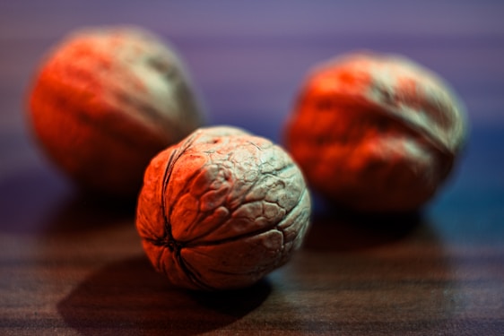 Three walnuts are placed on a wooden surface with a shallow depth of field. The focus is on the walnut in the foreground, highlighting its rugged texture and natural patterns.