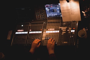 A person is operating a professional audio mixing console. The console features multiple faders, knobs, and illuminated buttons, alongside digital displays showing various audio settings. A white sheet of paper with text is placed on the right side of the console. The lighting is dim, casting a warm glow over the equipment.