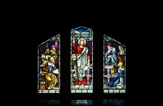 A stained glass window with three panels, featuring religious figures in vibrant colors. The central figure is depicted with a raised arm, surrounded by other figures engaged in various activities.