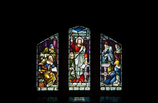 A stained glass window with three panels, featuring religious figures in vibrant colors. The central figure is depicted with a raised arm, surrounded by other figures engaged in various activities.