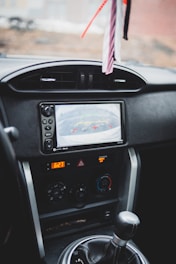 A car's interior dashboard featuring a rear-view camera display, various control knobs for air conditioning, and a digital clock showing the time 6:27. Hanging decor can be seen above the dashboard.
