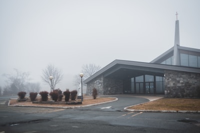 A foggy scene with a modern church building featuring a stone facade and large glass windows, situated on a quiet, empty parking lot. Sparse landscaping includes leafless trees and trimmed bushes, contributing to a serene and slightly eerie atmosphere.