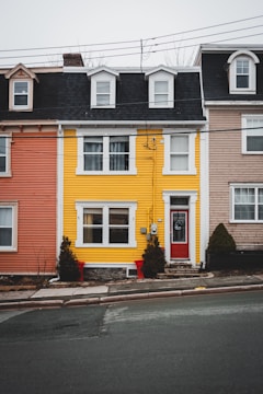 A row of colorful townhouses is aligned along a city street. The central house is striking with its bright yellow exterior and contrasting red door. On either side, the houses are painted in muted shades of pink and beige. The houses feature multiple windows with white frames and black gabled roofs. Small shrubs decorate the area in front of the yellow house.