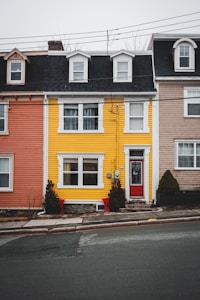 A row of colorful townhouses is aligned along a city street. The central house is striking with its bright yellow exterior and contrasting red door. On either side, the houses are painted in muted shades of pink and beige. The houses feature multiple windows with white frames and black gabled roofs. Small shrubs decorate the area in front of the yellow house.