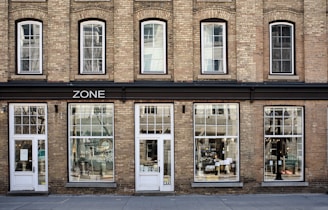 A brick building facade with large windows and multiple doors, featuring the word 'ZONE' above the entrance. The windows display reflections and various interior decorations, suggesting it might be a store. The building has an old-style architectural design with arched windows on the upper floor.