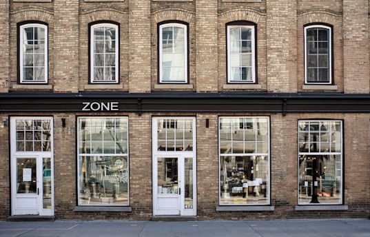 A brick building facade with large windows and multiple doors, featuring the word 'ZONE' above the entrance. The windows display reflections and various interior decorations, suggesting it might be a store. The building has an old-style architectural design with arched windows on the upper floor.