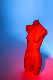 A red female torso mannequin stands against a gradient background transitioning from blue at the top to pinkish near the bottom. The lighting casts soft shadows and highlights the contours and curves of the mannequin.
