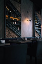 A dimly lit, modern restaurant interior featuring stacked firewood and book-lined shelves. The wall includes geometric wooden paneling and an industrial-style light fixture casting a warm glow. Tables are set with utensils and napkin holders, creating an inviting but intimate atmosphere.