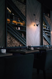 A dimly lit, modern restaurant interior featuring stacked firewood and book-lined shelves. The wall includes geometric wooden paneling and an industrial-style light fixture casting a warm glow. Tables are set with utensils and napkin holders, creating an inviting but intimate atmosphere.