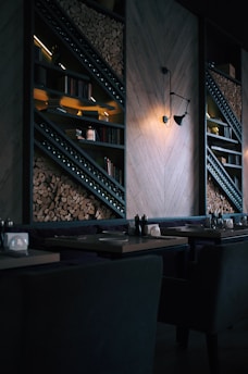 A dimly lit, modern restaurant interior featuring stacked firewood and book-lined shelves. The wall includes geometric wooden paneling and an industrial-style light fixture casting a warm glow. Tables are set with utensils and napkin holders, creating an inviting but intimate atmosphere.