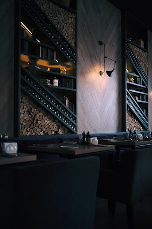 A dimly lit, modern restaurant interior featuring stacked firewood and book-lined shelves. The wall includes geometric wooden paneling and an industrial-style light fixture casting a warm glow. Tables are set with utensils and napkin holders, creating an inviting but intimate atmosphere.