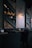 A dimly lit, modern restaurant interior featuring stacked firewood and book-lined shelves. The wall includes geometric wooden paneling and an industrial-style light fixture casting a warm glow. Tables are set with utensils and napkin holders, creating an inviting but intimate atmosphere.