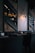 A dimly lit, modern restaurant interior featuring stacked firewood and book-lined shelves. The wall includes geometric wooden paneling and an industrial-style light fixture casting a warm glow. Tables are set with utensils and napkin holders, creating an inviting but intimate atmosphere.