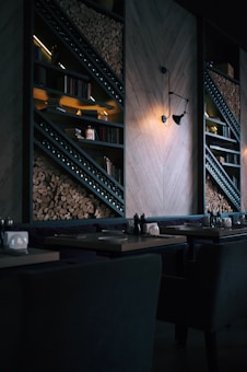 A dimly lit, modern restaurant interior featuring stacked firewood and book-lined shelves. The wall includes geometric wooden paneling and an industrial-style light fixture casting a warm glow. Tables are set with utensils and napkin holders, creating an inviting but intimate atmosphere.