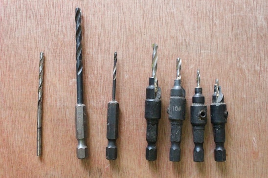 Seven drill bits of varying sizes are lined up on a wooden surface. The bits have a metallic sheen and appear to be well-used, with some showing signs of wear.