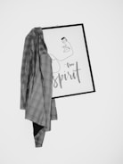 A minimalist artwork with a one-line drawing of a face and the words 'free spirit' is partially covered by a plaid blazer. The art is framed in a simple black frame and hangs against a plain white wall.