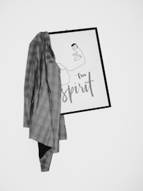 A minimalist artwork with a one-line drawing of a face and the words 'free spirit' is partially covered by a plaid blazer. The art is framed in a simple black frame and hangs against a plain white wall.