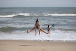 A child in a blue swimsuit is captured mid-air as they leap above the sandy beach, with the ocean waves in the background. Two other people, wearing swimsuits, are seen standing in the shallow water.