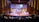 A large auditorium is filled with people, with a focus on a stage where a person is speaking at a podium. The stage is decorated with large puzzle pieces and a backdrop screen displaying text. Musicians are seated to the left side of the stage, and there is subtle lighting creating a warm atmosphere.