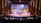 A large auditorium is filled with people, with a focus on a stage where a person is speaking at a podium. The stage is decorated with large puzzle pieces and a backdrop screen displaying text. Musicians are seated to the left side of the stage, and there is subtle lighting creating a warm atmosphere.