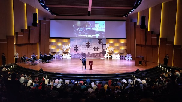 A large auditorium is filled with people, with a focus on a stage where a person is speaking at a podium. The stage is decorated with large puzzle pieces and a backdrop screen displaying text. Musicians are seated to the left side of the stage, and there is subtle lighting creating a warm atmosphere.