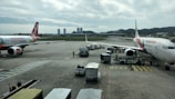 Airplanes are parked on an airport tarmac with airport ground handling vehicles and personnel around them. One plane has a colorful livery promoting 'Sustainable ASEAN Tourism', while another displays airline branding. The scene includes overcast skies and distant background buildings.