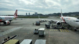 Airplanes are parked on an airport tarmac with airport ground handling vehicles and personnel around them. One plane has a colorful livery promoting 'Sustainable ASEAN Tourism', while another displays airline branding. The scene includes overcast skies and distant background buildings.