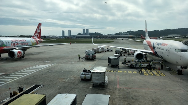 Airplanes are parked on an airport tarmac with airport ground handling vehicles and personnel around them. One plane has a colorful livery promoting 'Sustainable ASEAN Tourism', while another displays airline branding. The scene includes overcast skies and distant background buildings.