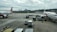 Airplanes are parked on an airport tarmac with airport ground handling vehicles and personnel around them. One plane has a colorful livery promoting 'Sustainable ASEAN Tourism', while another displays airline branding. The scene includes overcast skies and distant background buildings.