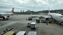 Airplanes are parked on an airport tarmac with airport ground handling vehicles and personnel around them. One plane has a colorful livery promoting 'Sustainable ASEAN Tourism', while another displays airline branding. The scene includes overcast skies and distant background buildings.