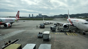 Airplanes are parked on an airport tarmac with airport ground handling vehicles and personnel around them. One plane has a colorful livery promoting 'Sustainable ASEAN Tourism', while another displays airline branding. The scene includes overcast skies and distant background buildings.