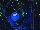 A close-up view of a computer's internal hardware components, including a central processing unit (CPU) cooler with a prominently displayed Intel logo. The lighting has a blue tone, highlighting the intricacy of the various cables, circuit boards, and heat sinks in the assembly.