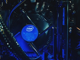 A close-up view of a computer's internal hardware components, including a central processing unit (CPU) cooler with a prominently displayed Intel logo. The lighting has a blue tone, highlighting the intricacy of the various cables, circuit boards, and heat sinks in the assembly.